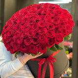 Large hat box bouquet of red roses in a black box with a red ribbon, held by a person.